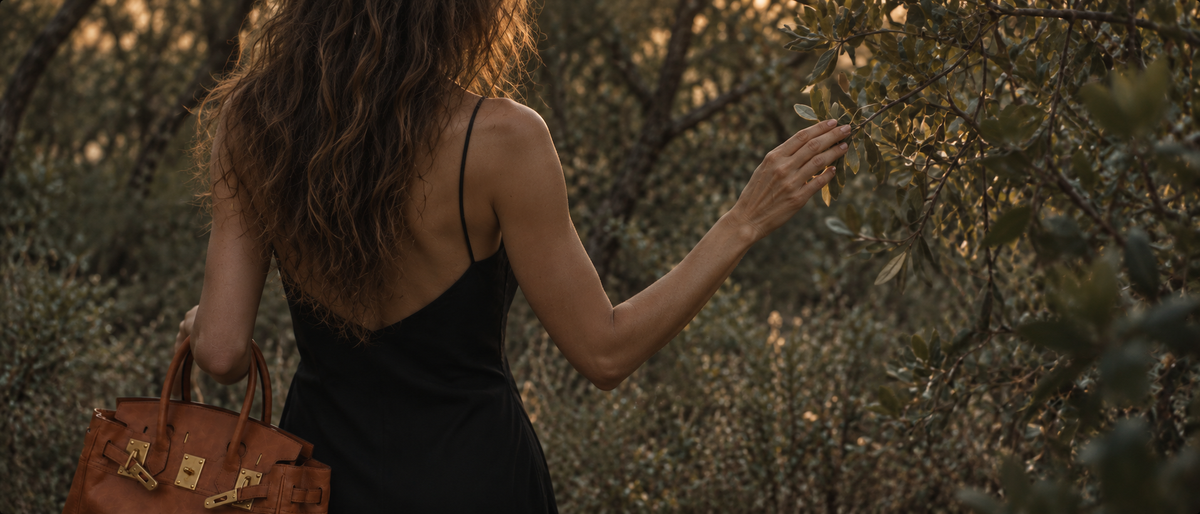 Woman in a black dress carrying the Cleo dark orange leather tote among olive trees