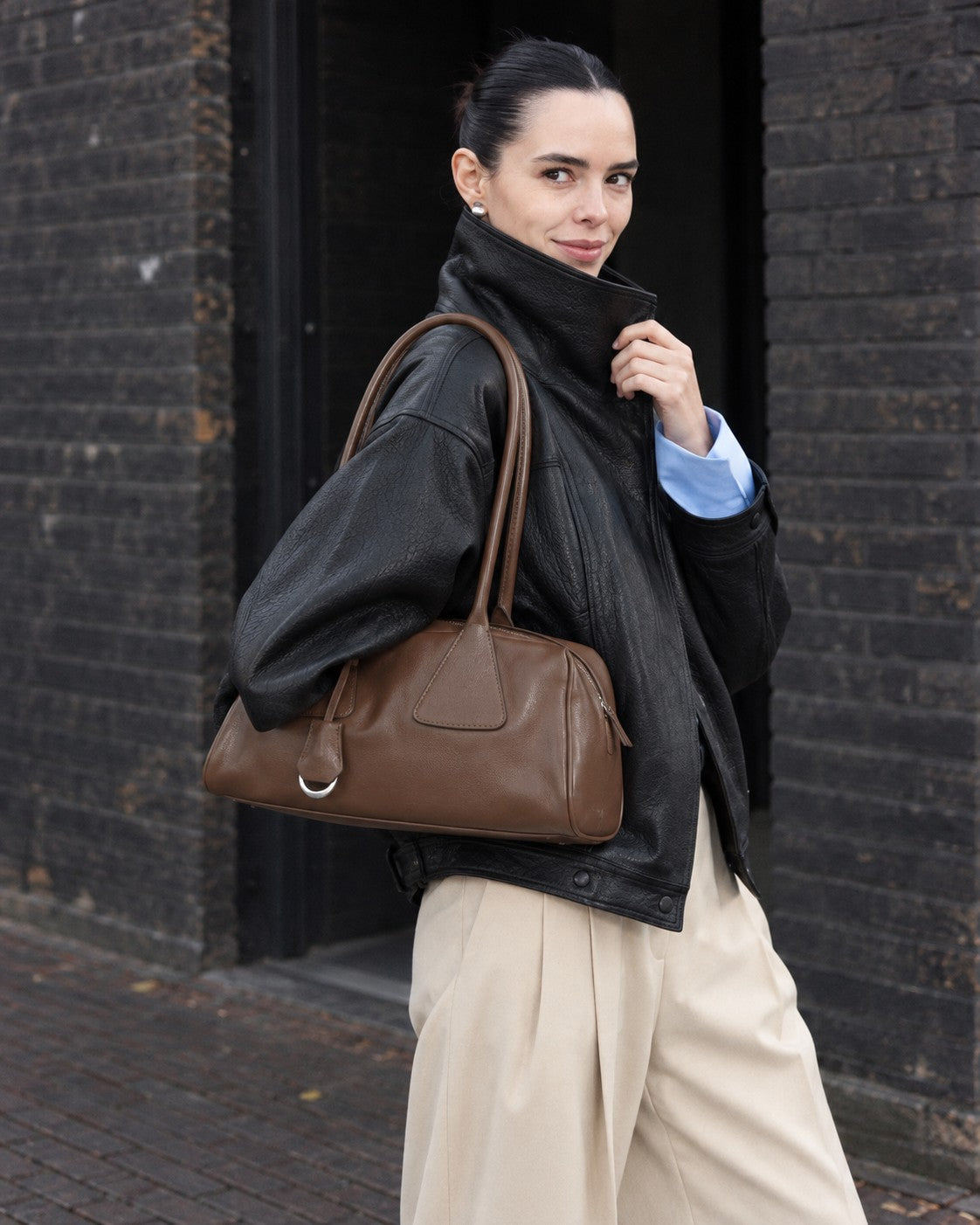 An adult model carrying the Bagulia Vera shoulder bag in brown on a brick-lined street.