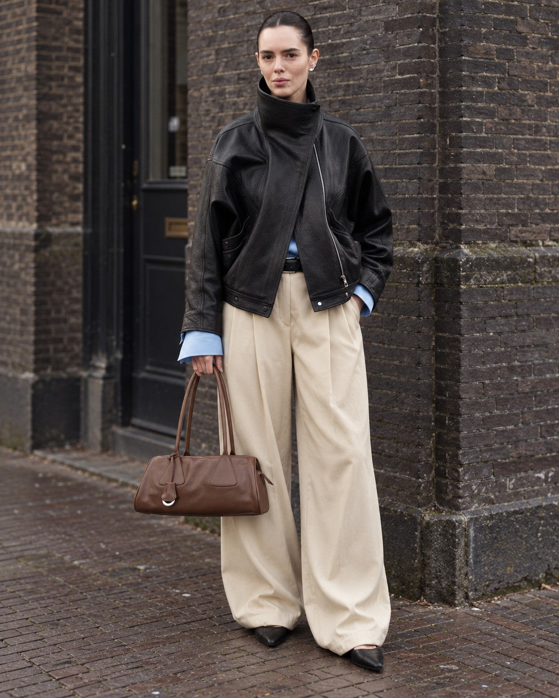 An adult model carrying the Bagulia Vera shoulder bag in brown on a brick-lined street.