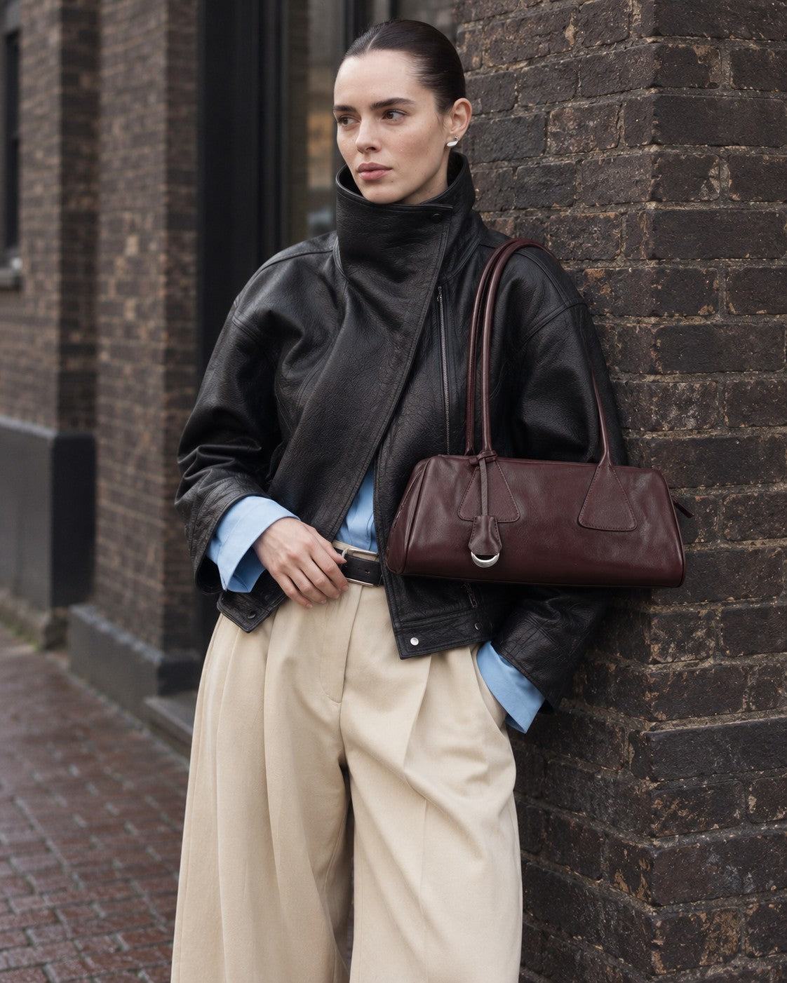 An adult model carrying the Bagulia Vera shoulder bag in Oxblood on a brick-lined street.