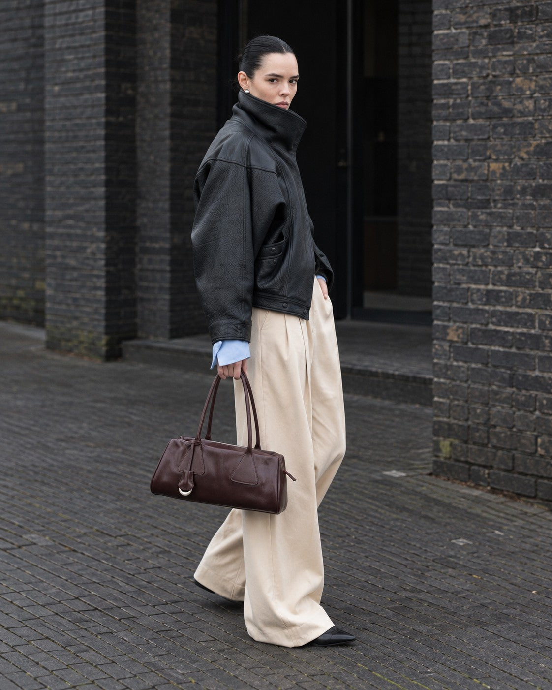 An adult model carrying the Bagulia Vera shoulder bag in Oxblood on a brick-lined street.